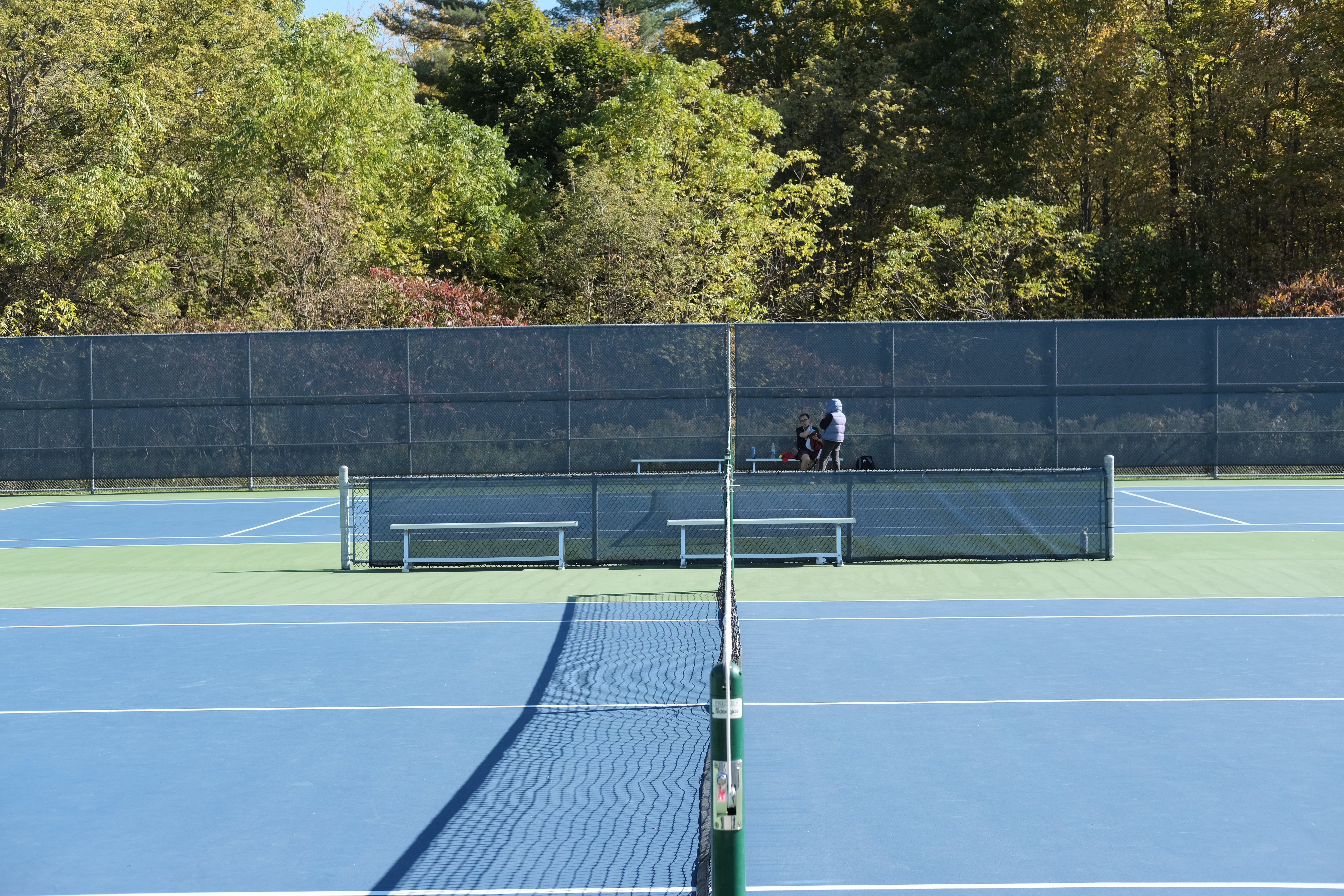 Two outdoor blue and green acrylic hardcourts at the University of Toronto Tennis Centre in Scarborough, with players on the far side and trees in the background.